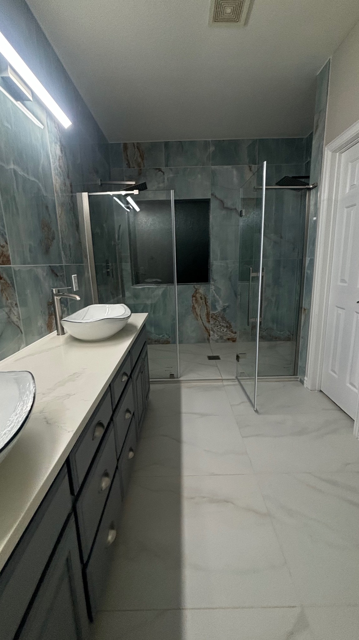 Nowoczesna łazienka z kabiną prysznicową typu walk-in, umywalkami nablatowymi i marmurowymi płytkami w odcieniach zieleni i bieli. Minimalistyczny design, eleganckie wykończenie.