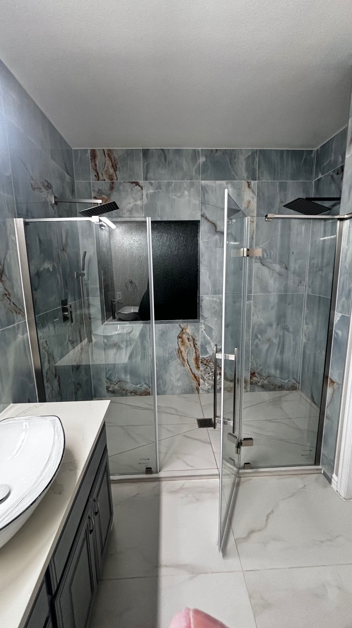 Nowoczesna łazienka z kabiną prysznicową typu walk-in, wykończona płytkami imitującymi marmur w odcieniach szarości i brązu. Widoczna umywalka nablatowa i czarna wnęka.