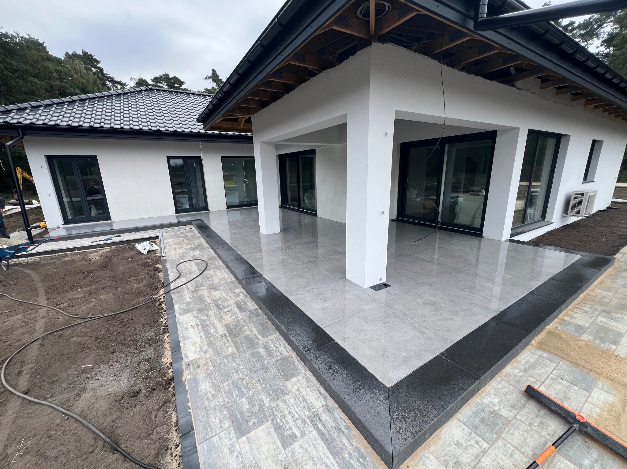 Nowoczesny taras z szarych płyt i obrzeżem z czarnej kostki, przylegający do białego domu z czarną stolarką okienną. Częściowo ułożona kostka brukowa na podjeździe.