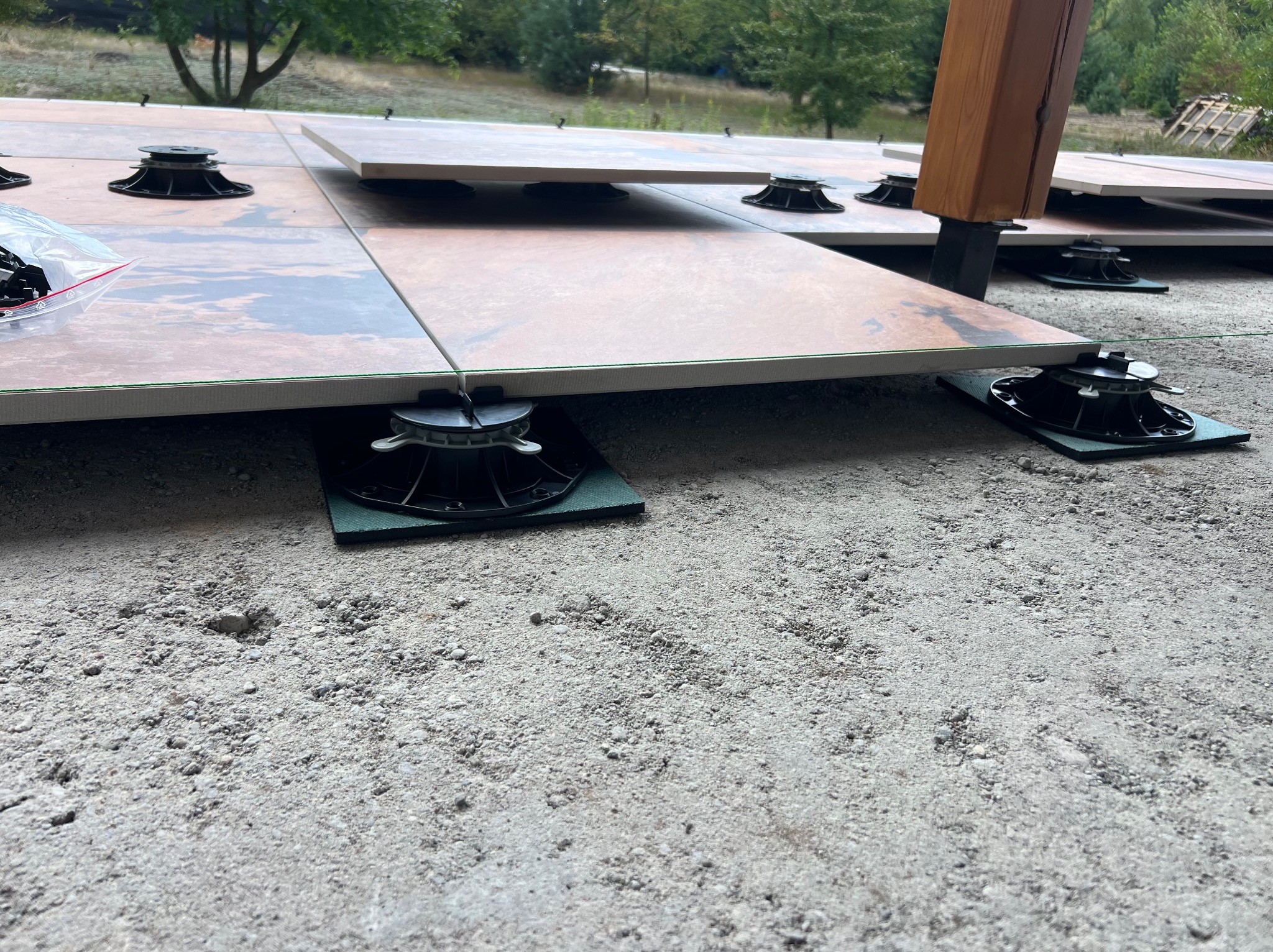 Montaż podniesionej podłogi tarasowej z płytek na regulowanych wspornikach. Widoczne elementy mocujące i podłoże z kruszywa. Częściowo ułożone płyty tarasowe.