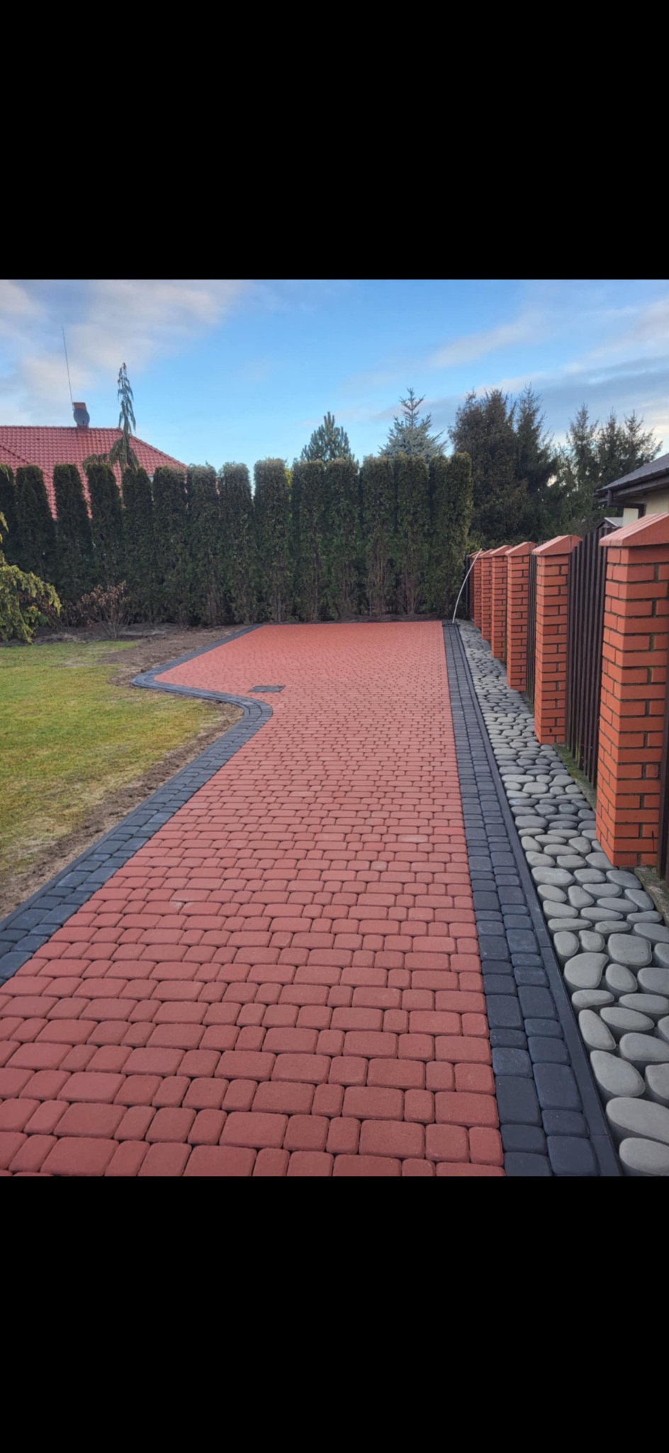Czerwona kostka brukowa ułożona na podjeździe, obrzeża z ciemnej kostki i kamieni, ogrodzenie z cegły i metalu, wysokie tuje w tle, błękitne niebo.