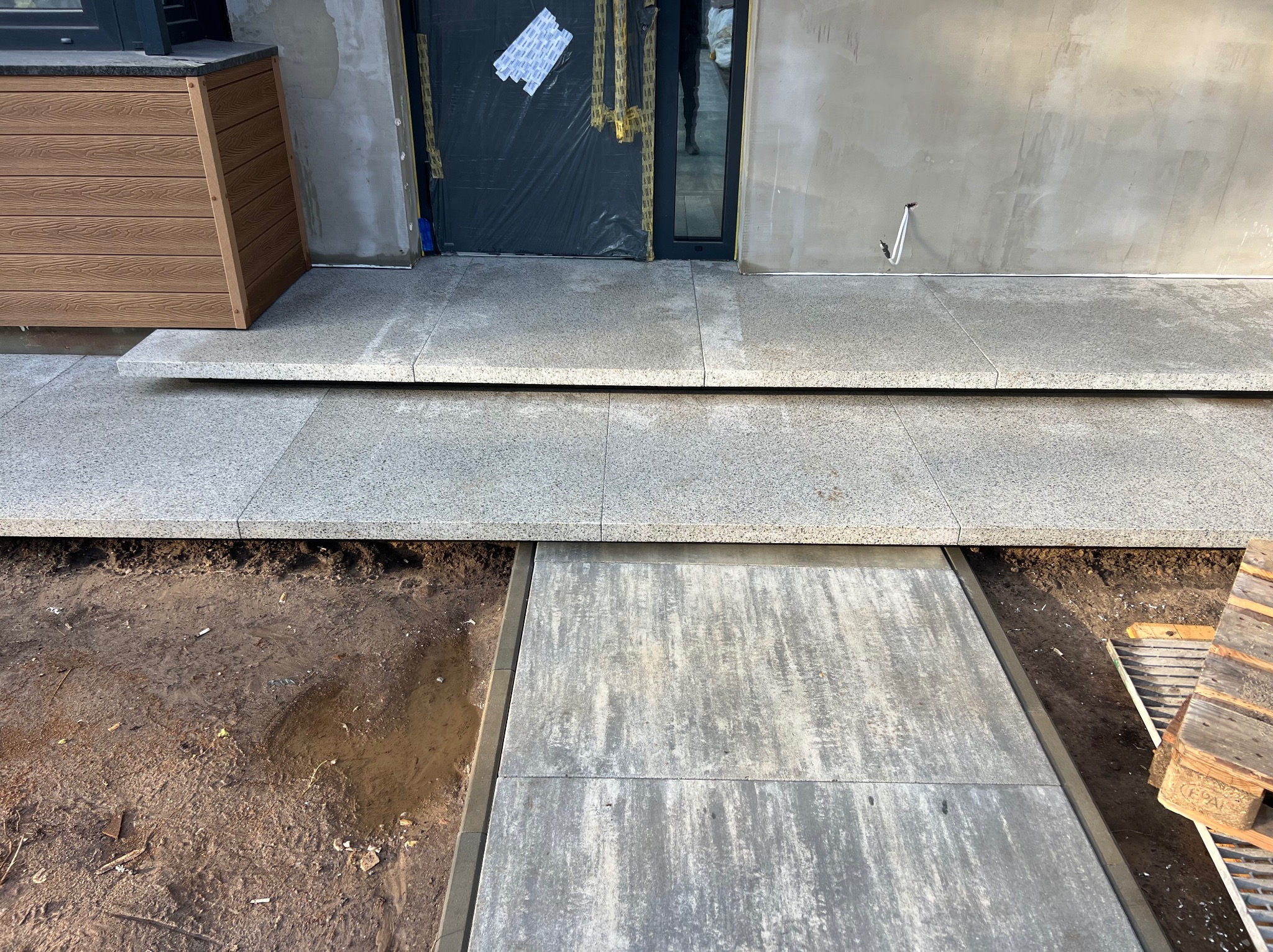 Granitowa nawierzchnia tarasu z dwoma stopniami, widok z góry na fragment ziemi i paletę. Drzwi wejściowe zabezpieczone folią. Nowoczesne wykończenie.