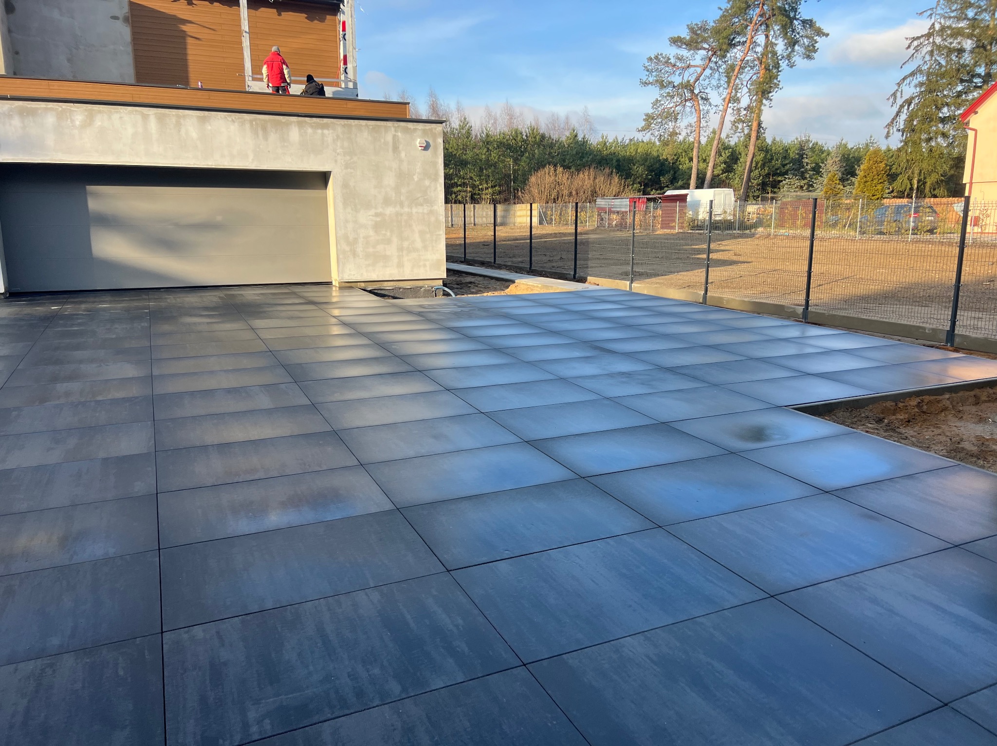 Nowoczesny podjazd z dużych, grafitowych płyt betonowych, częściowo ułożonych, z widocznym przygotowaniem podłoża. W tle dom z garażem i ogrodzenie.