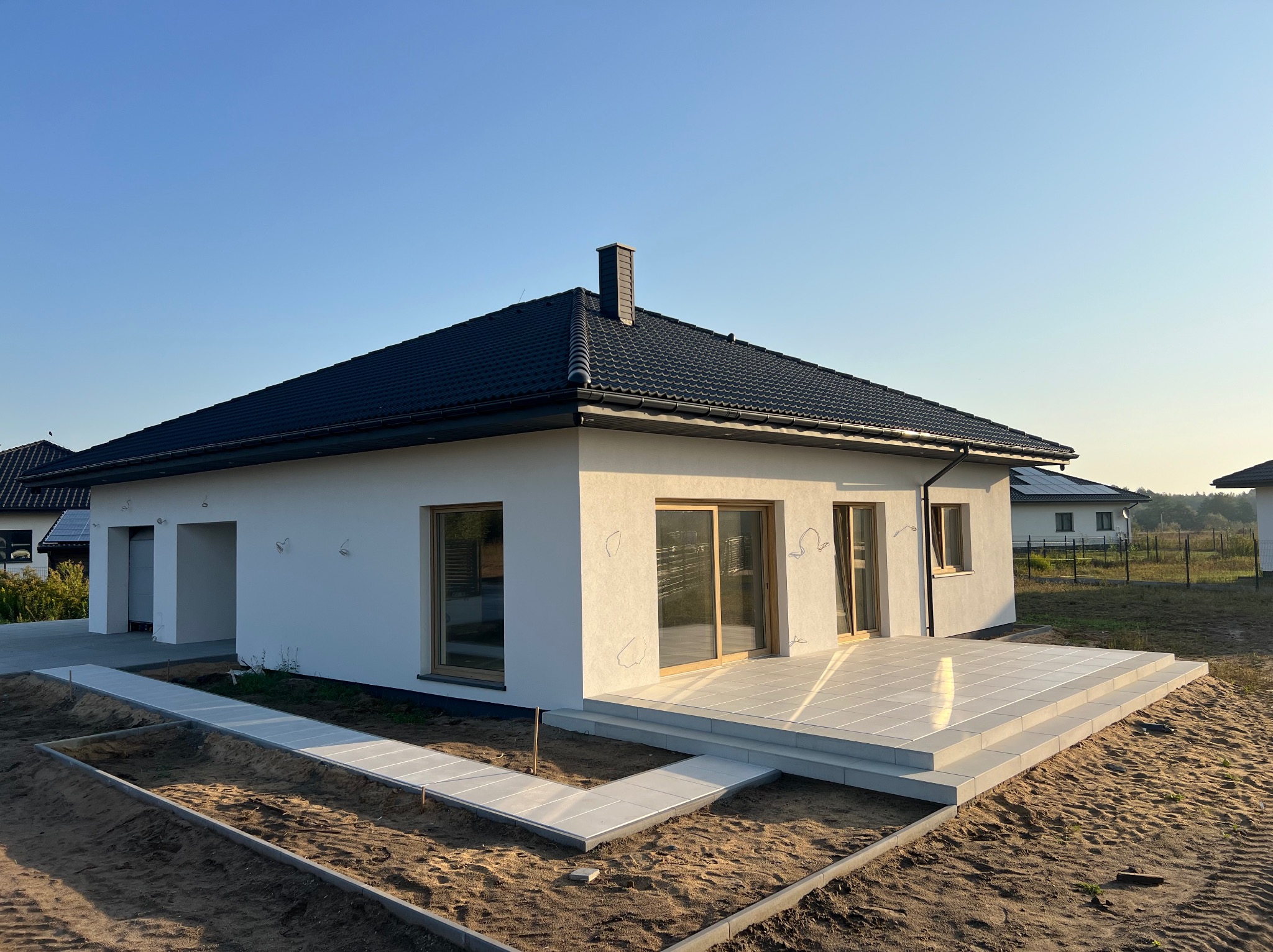 Nowoczesny dom z białymi ścianami i tarasem z jasnych płyt. Ułożona ścieżka z kostki prowadzi do domu. W tle błękitne niebo i inne domy.