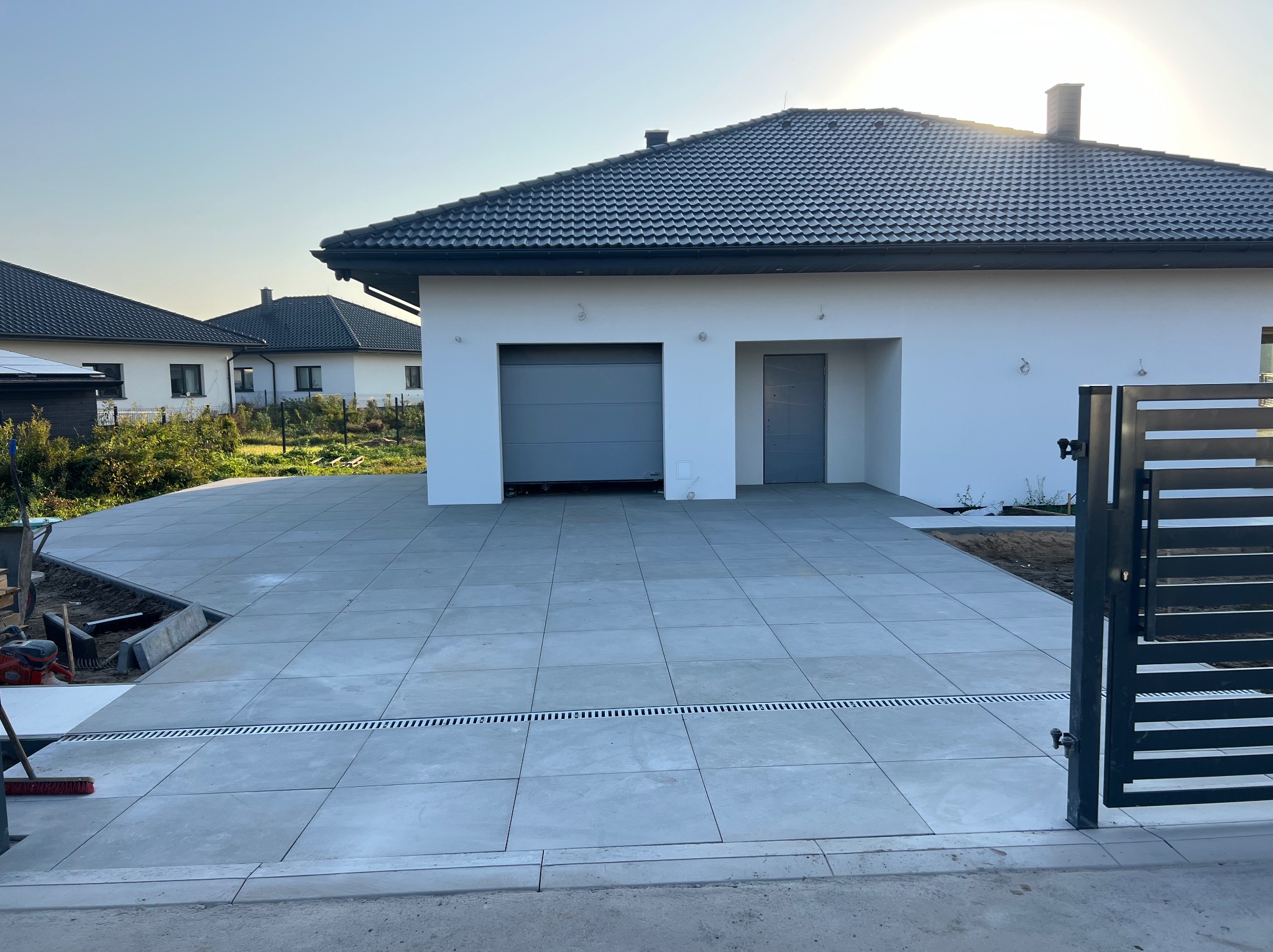 Nowoczesny podjazd z szarych płyt betonowych prowadzący do garażu, z widocznym systemem odprowadzania wody i fragmentem otwartej bramy w tle.