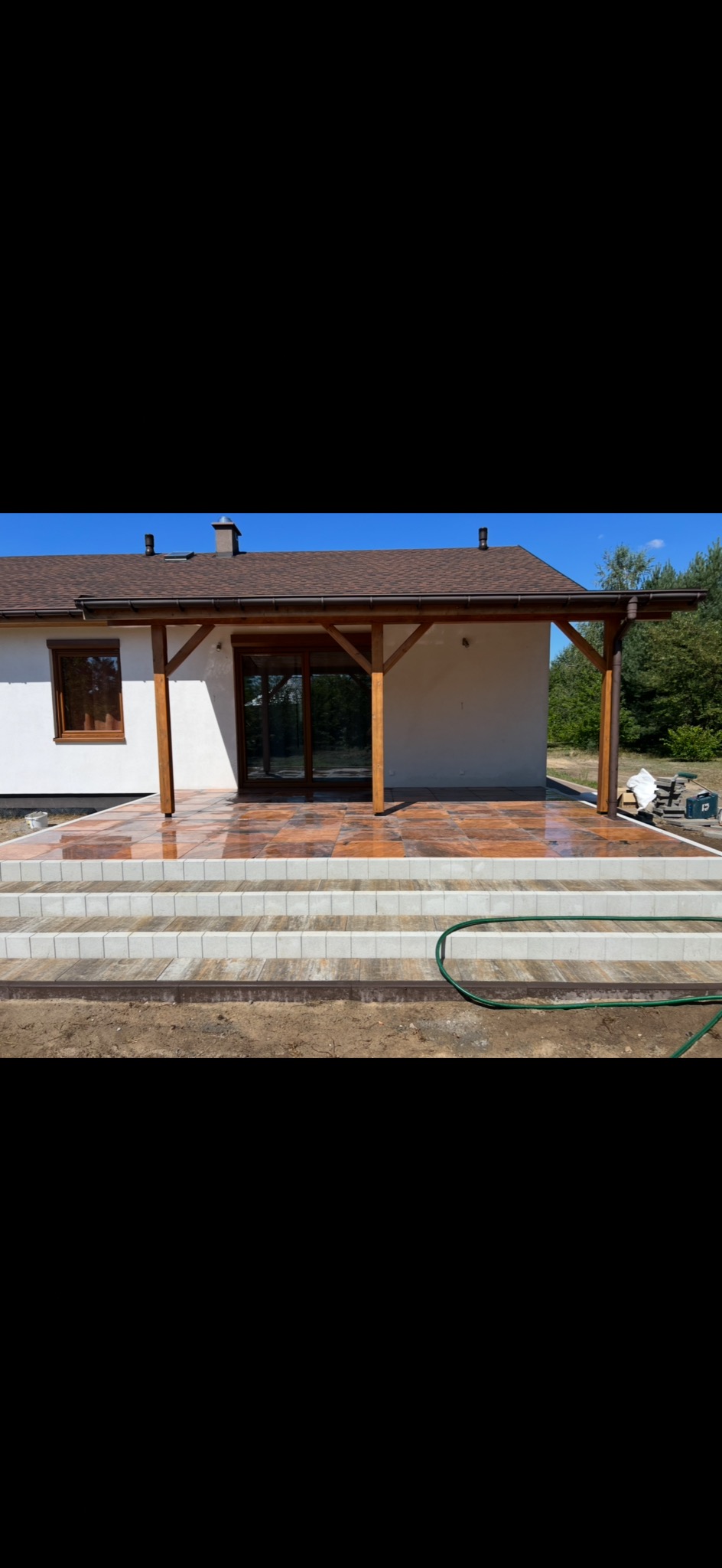 Błyszczący taras z kostki brukowej w odcieniach brązu, prowadzący do domu z drewnianą pergolą. Widoczne schody z jasnej kostki i fragment węża ogrodowego.