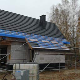 Hubert Siepiatowski - Budowa domu jednorodzinnego w trakcie krycia dachu czarną dachówką. Widoczny dekarz na dachu, rusztowania i częściowo ułożona więźba. Jesienna sceneria w tle.