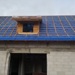 Hubert Siepiatowski - Wiązar dachowy z membraną w kolorze niebieskim, widok na okno dachowe w Płońsku. Konstrukcja dachu w trakcie budowy, ściany z białego bloczka.