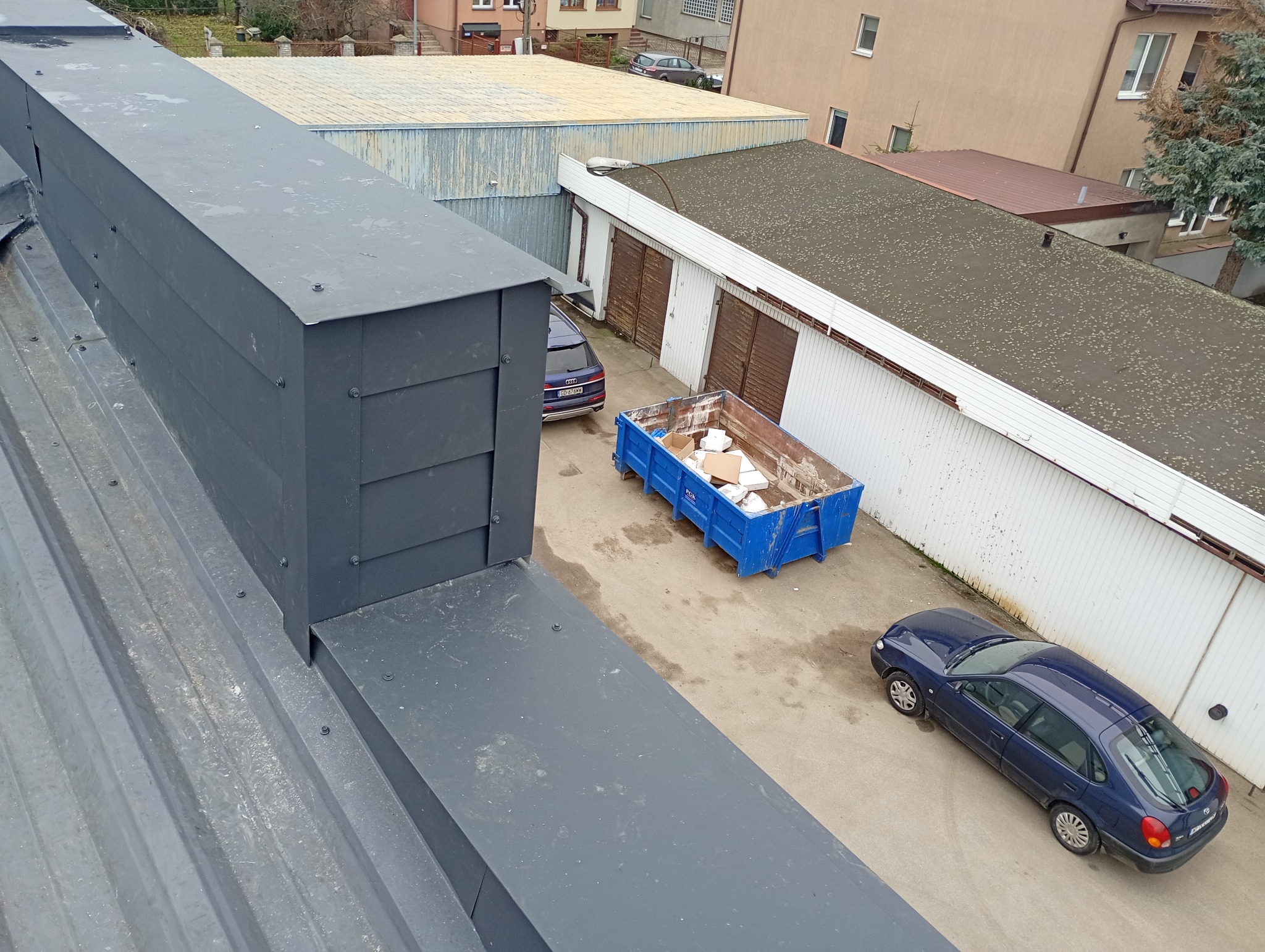 Szary, metalowy dach z kominem wentylacyjnym. W tle budynki gospodarcze, kontener na odpady i dwa samochody. Widok z góry, z bliska na fragment dachu.