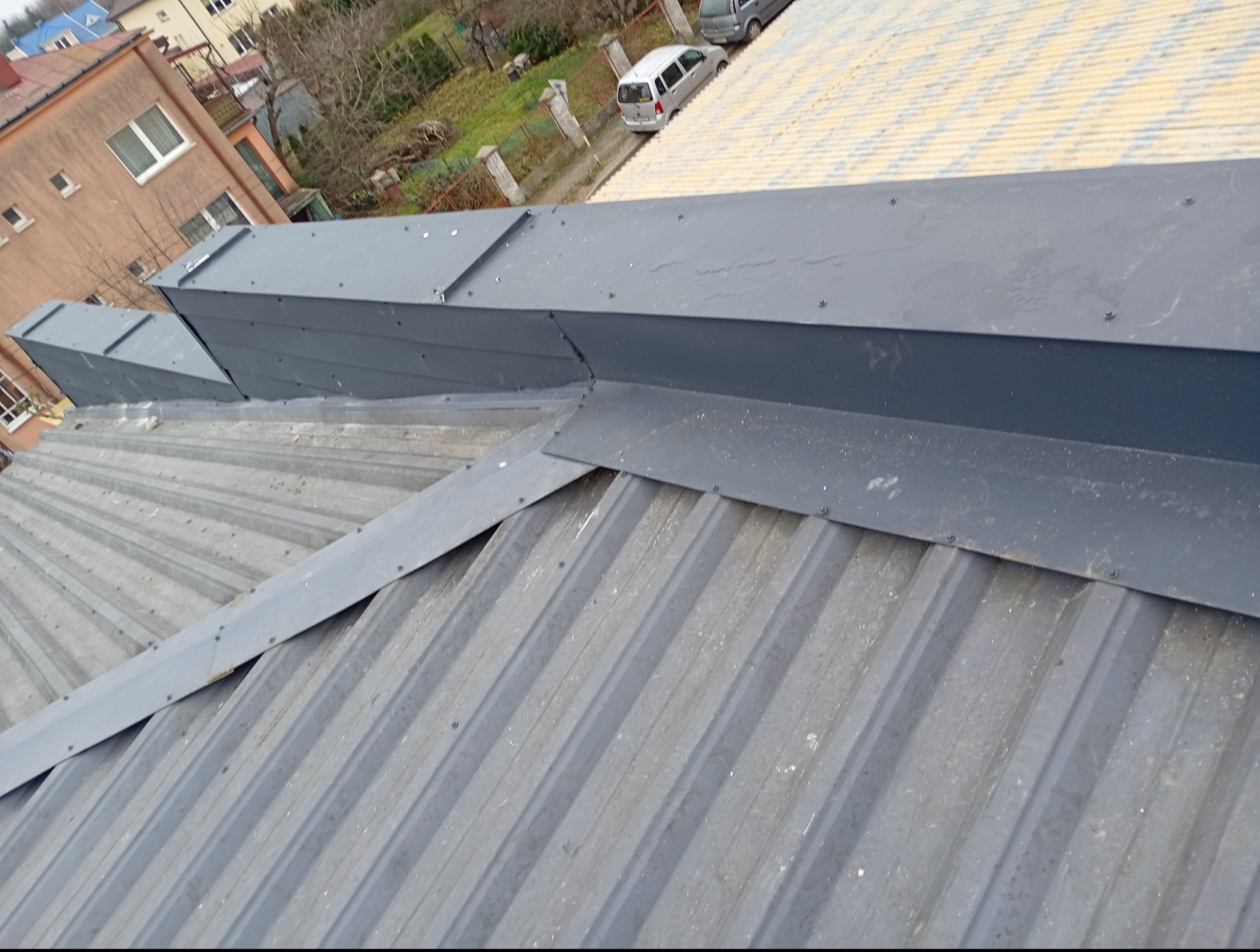 Szary dach z blachy trapezowej z nowymi obróbkami blacharskimi. Widok z góry, na tle zabudowań i drzew.