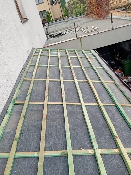 Wstępny etap budowy dachu płaskiego z widoczną konstrukcją drewnianą na czarnej membranie, z fragmentem budynku w tle i zielonymi drzewami w górnej części kadru.