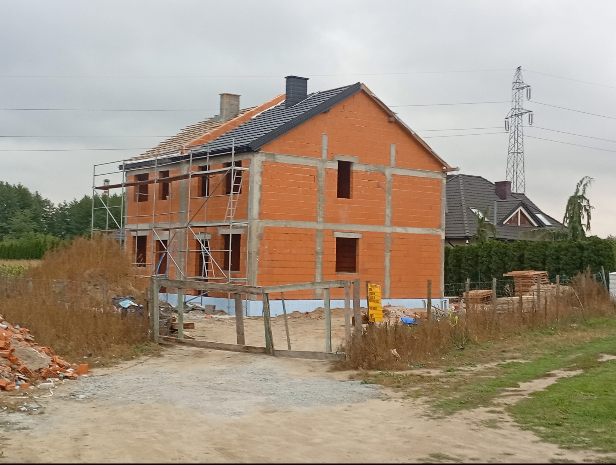 Budowa domu z częściowo ukończonym dachem z czarnej dachówki, widoczne rusztowania i materiały budowlane. W tle słup wysokiego napięcia i zieleń.