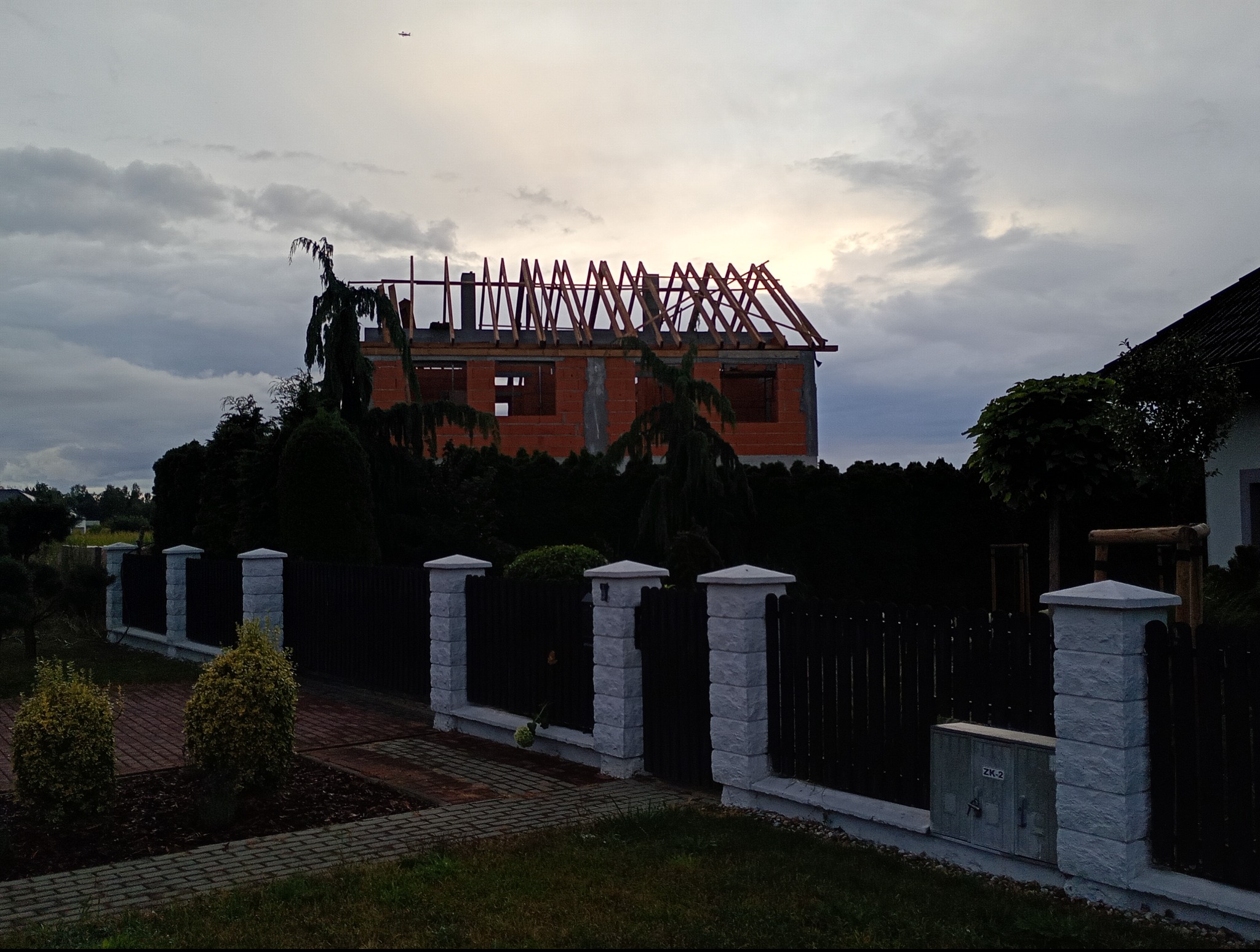 Budowa domu jednorodzinnego w trakcie realizacji: widoczna konstrukcja więźby dachowej na tle pochmurnego nieba. Ujęcie zza ogrodzenia.