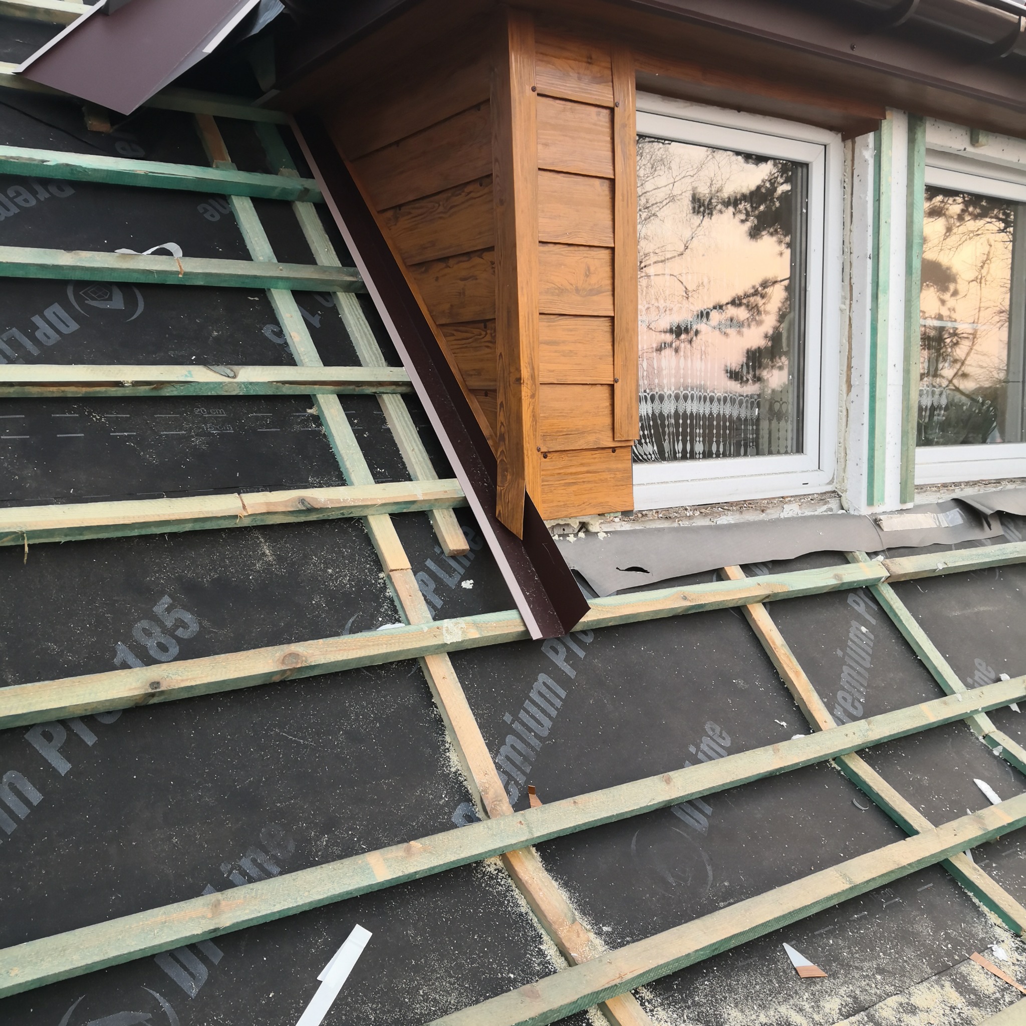 W trakcie prac dekarskich: widok na drewnianą konstrukcję dachu pokrytą membraną, z elementami obróbki blacharskiej przy oknie. Widoczne detale drewnianej elewacji.