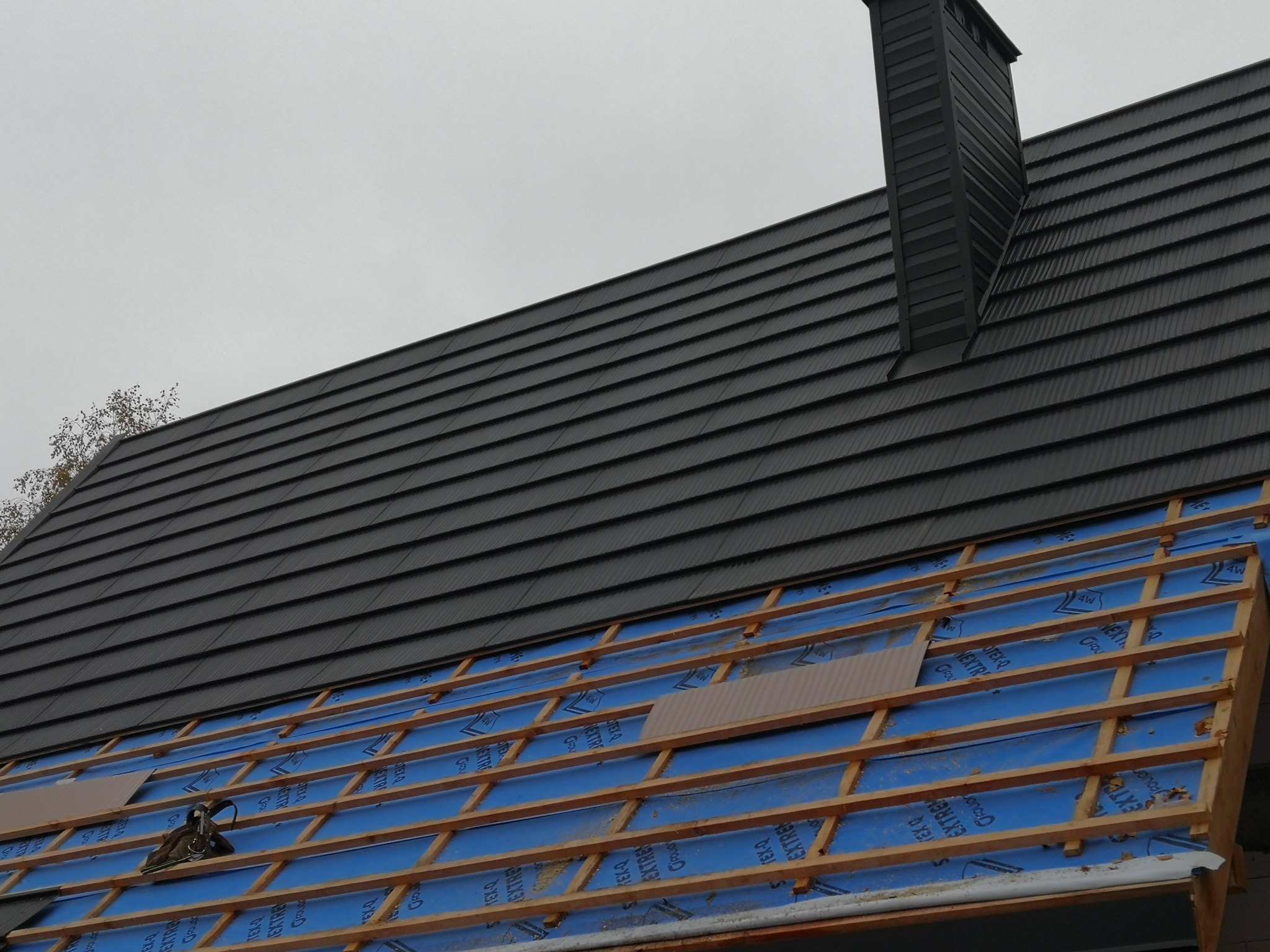 Częściowo pokryty dach z czarną dachówką, widoczna więźba dachowa z niebieską membraną i komin. W tle pochmurne niebo.