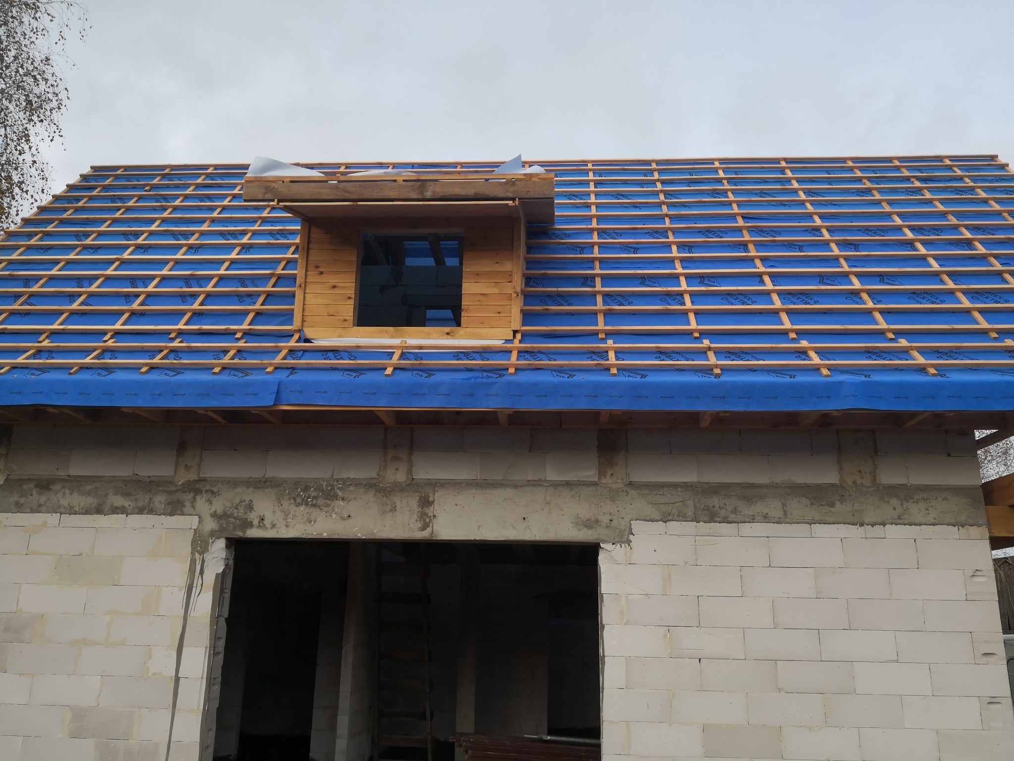 Wiązar dachowy z membraną w kolorze niebieskim, widok na okno dachowe w Płońsku. Konstrukcja dachu w trakcie budowy, ściany z białego bloczka.