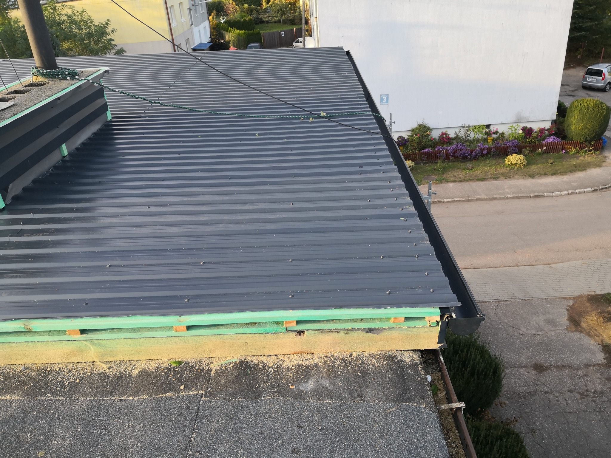 Szary dach z blachy trapezowej, widok z góry, z widocznym ociepleniem krawędzi dachu i rynną. W tle budynki i zieleń.