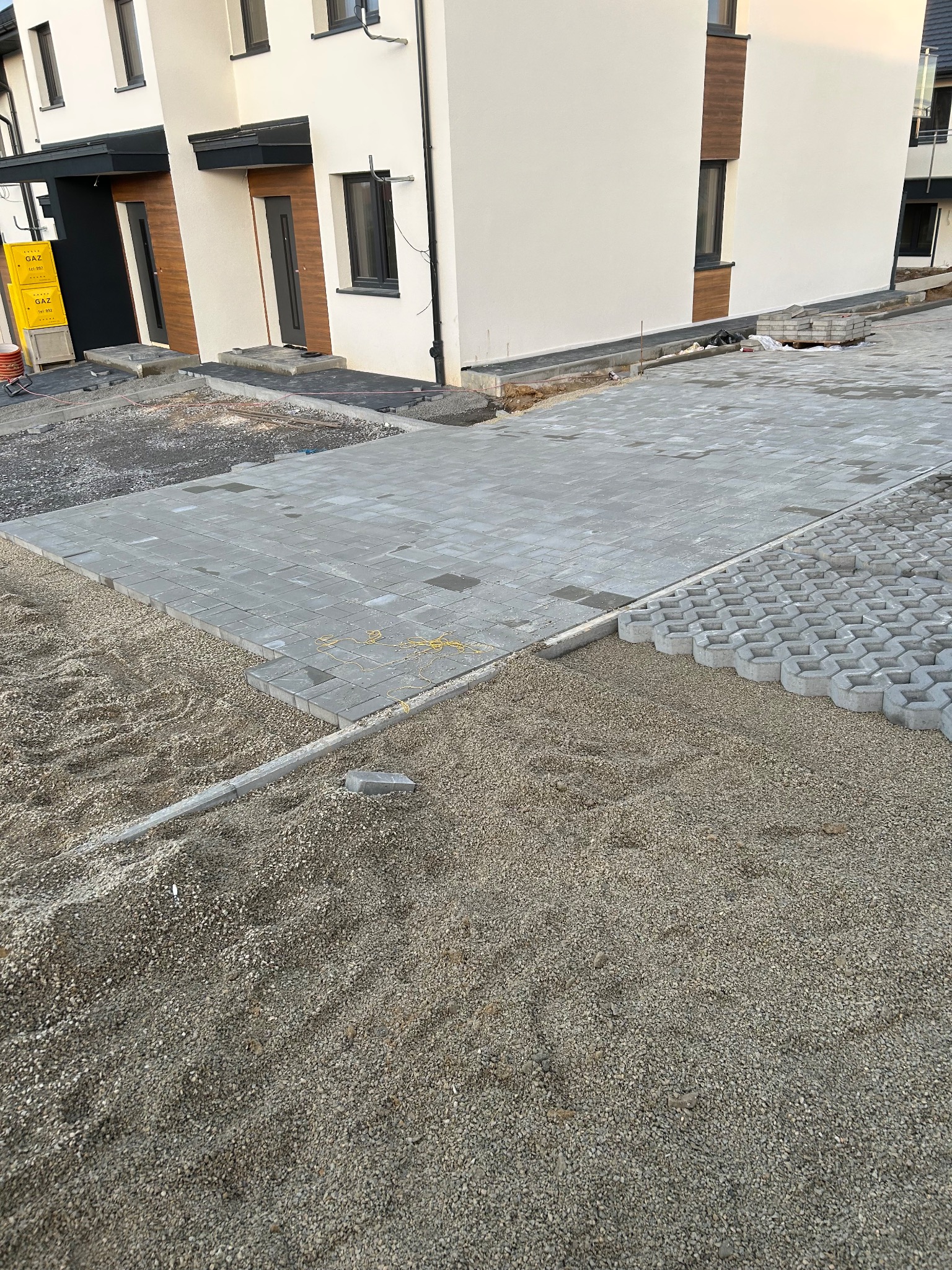 Układanie szarej kostki brukowej na podsypce żwirowej przed budynkiem. Częściowo ułożona nawierzchnia z kostki ażurowej. Widoczne krawężniki i przygotowane podłoże.