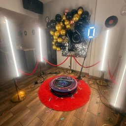 PleneroVo Events - Fotobudka 360 na czerwonym dywanie, otoczona złotymi słupkami z czerwonym sznurem. W tle dekoracja z balonów i błyszcząca ścianka. Konfetti na podłodze.