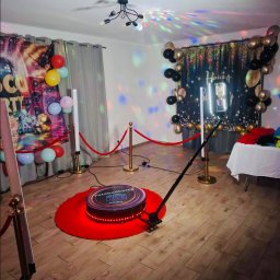 PleneroVo Events - Oświetlona platforma 360 fotobudki na czerwonym dywanie, otoczona złotymi słupkami i balonami, tworząca imprezową atmosferę w pomieszczeniu.