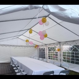 PleneroVo Events - Biały namiot z długim stołem i krzesłami, udekorowany balonami w kolorach różowym, białym i żółtym. Widok na zewnątrz przez okna namiotu.