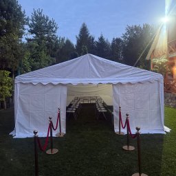 PleneroVo Events - Biały namiot eventowy na trawie, z rzędami krzeseł wewnątrz, oddzielony od wejścia złotymi słupkami z czerwonym sznurem. Tło stanowią drzewa i budynek.