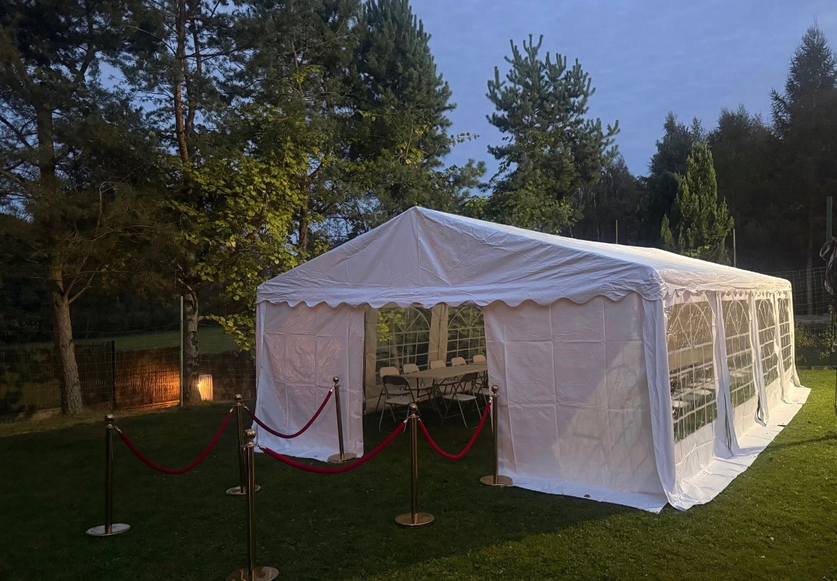 Biały namiot plenerowy z czerwonymi linami oddzielającymi wejście, ustawiony na trawie, otoczony drzewami. Wewnątrz widoczny stół i krzesła.
