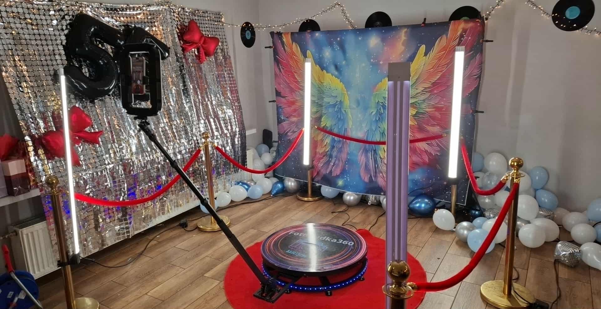 Ozdobiona przestrzeń z fotobudką 360, tłem z motywem skrzydeł, srebrną ścianką, balonami i czerwonymi sznurami, gotowa na imprezę. Widoczne oświetlenie LED.