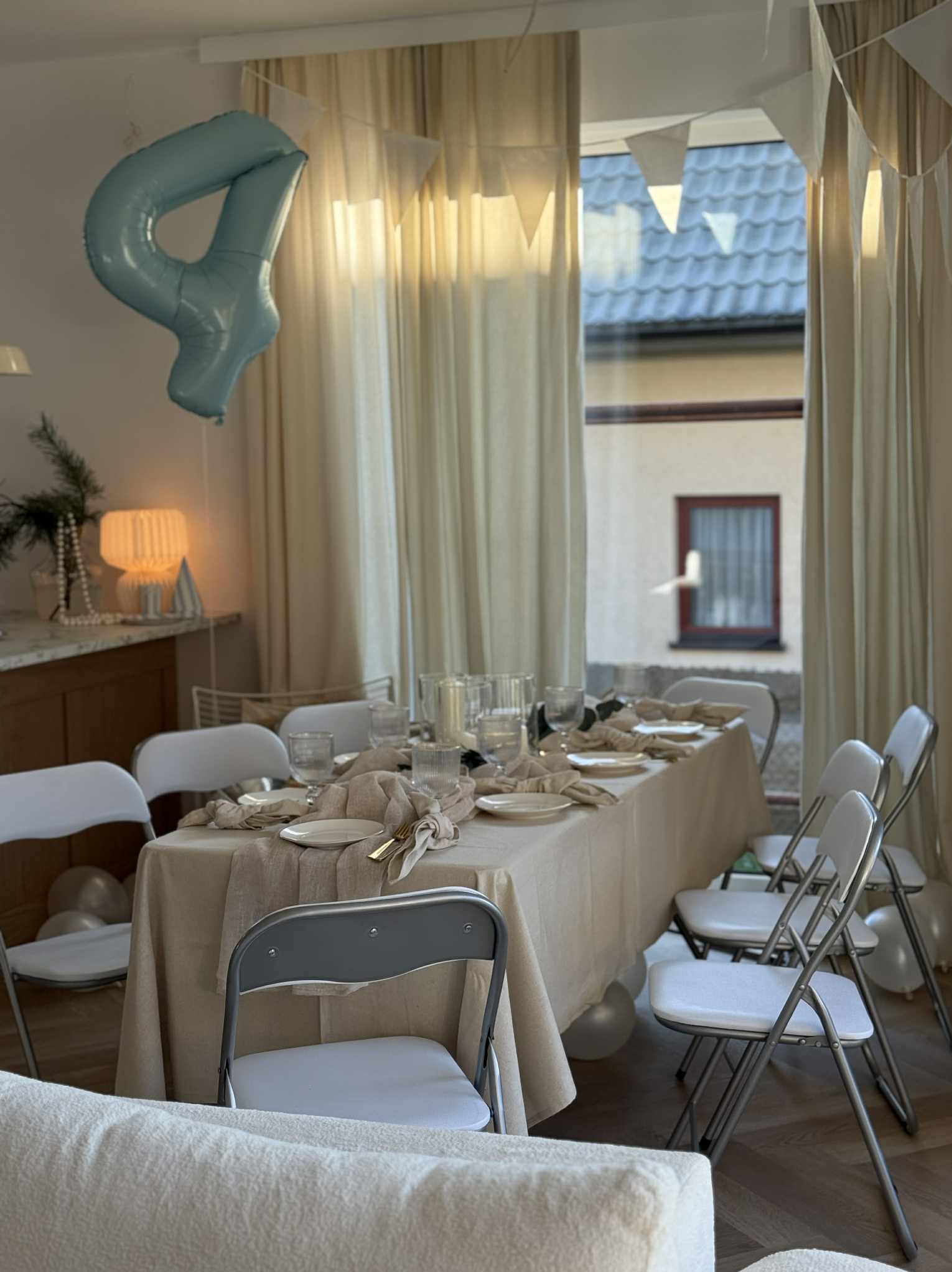 Uroczysty stół z beżowym obrusem, nakryty dla gości, z balonem w kształcie cyfry 4 i girlandą, ustawiony w jasnym wnętrzu z oknem.