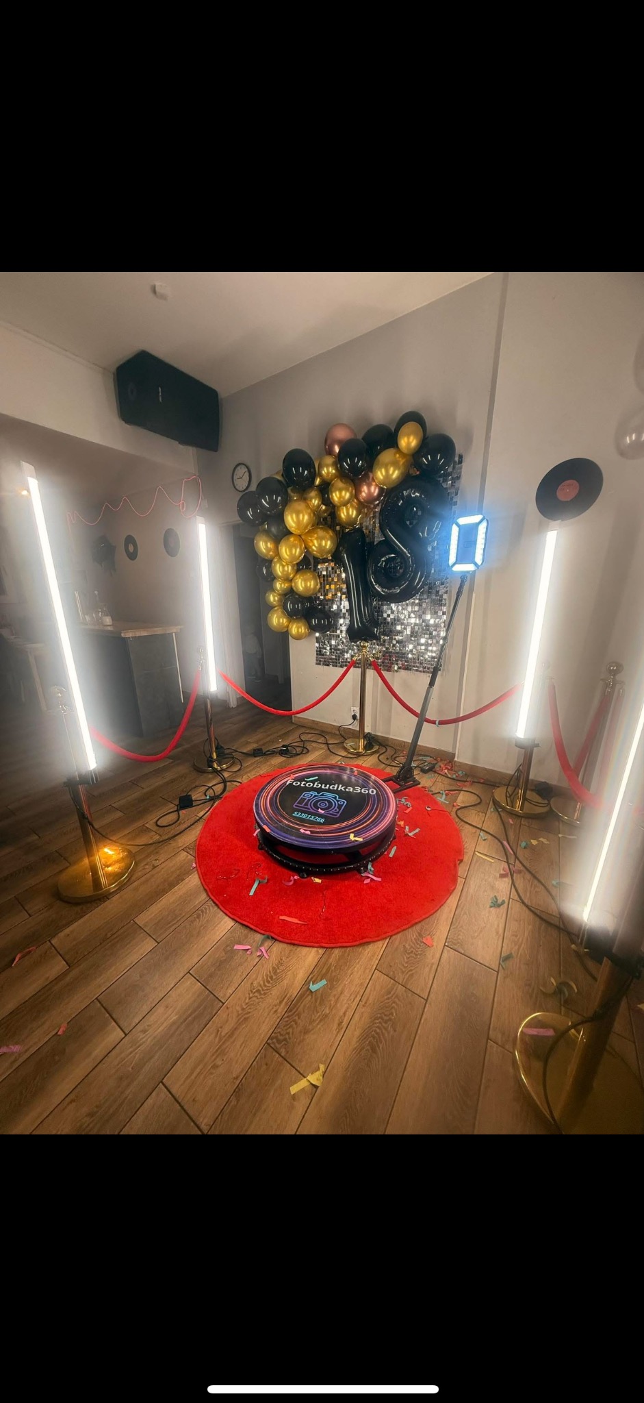 Fotobudka 360 na czerwonym dywanie, otoczona złotymi słupkami z czerwonym sznurem. W tle dekoracja z balonów i błyszcząca ścianka. Konfetti na podłodze.