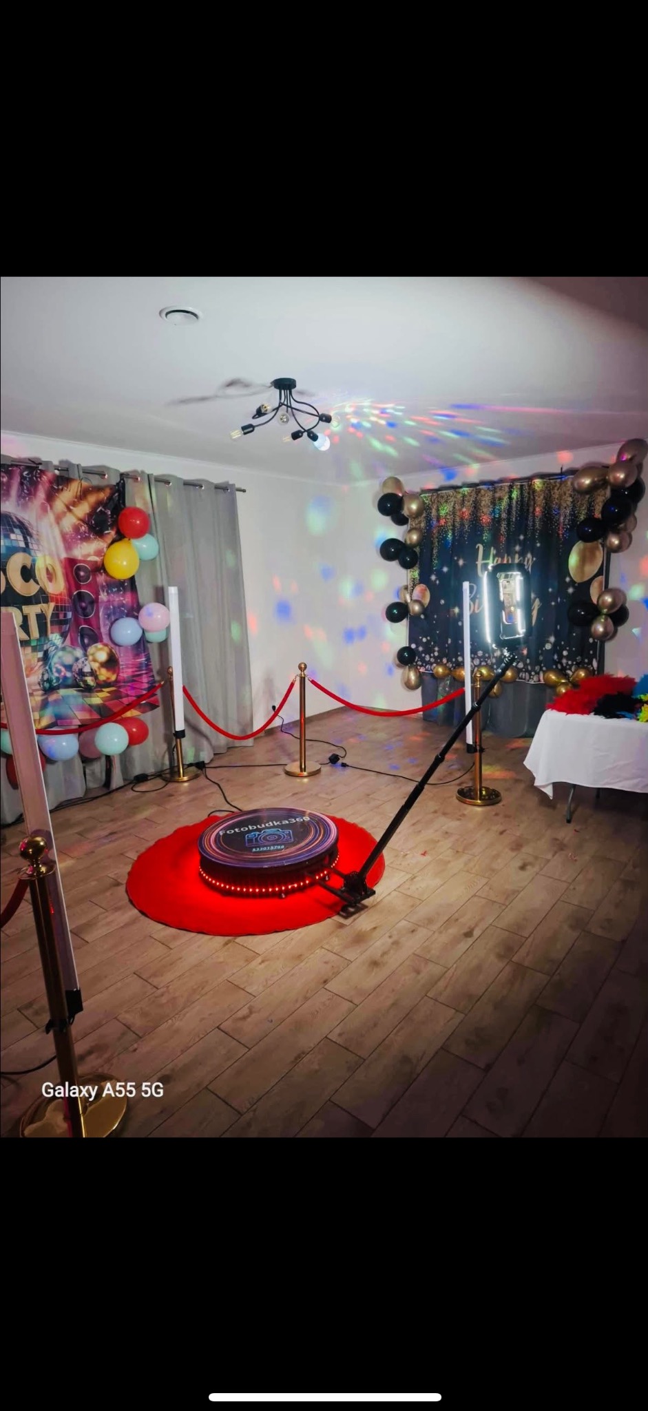 Oświetlona platforma 360 fotobudki na czerwonym dywanie, otoczona złotymi słupkami i balonami, tworząca imprezową atmosferę w pomieszczeniu.