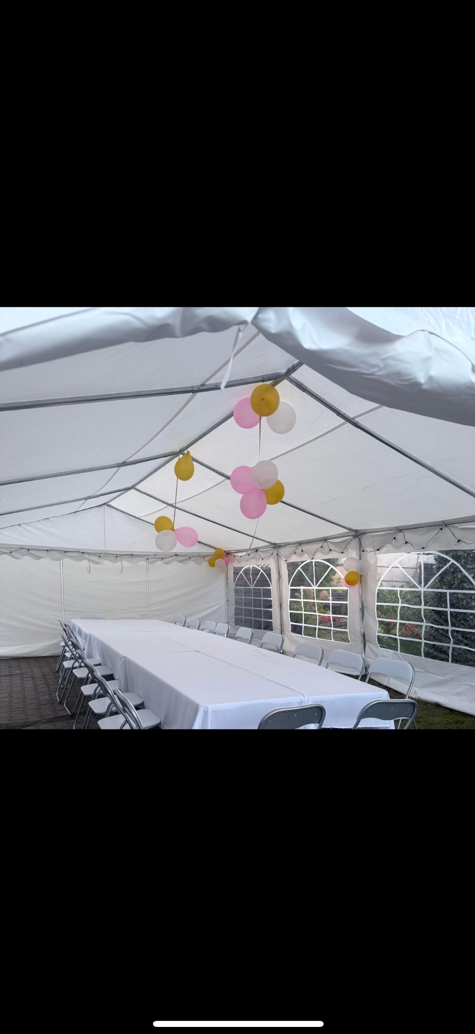 Biały namiot z długim stołem i krzesłami, udekorowany balonami w kolorach różowym, białym i żółtym. Widok na zewnątrz przez okna namiotu.