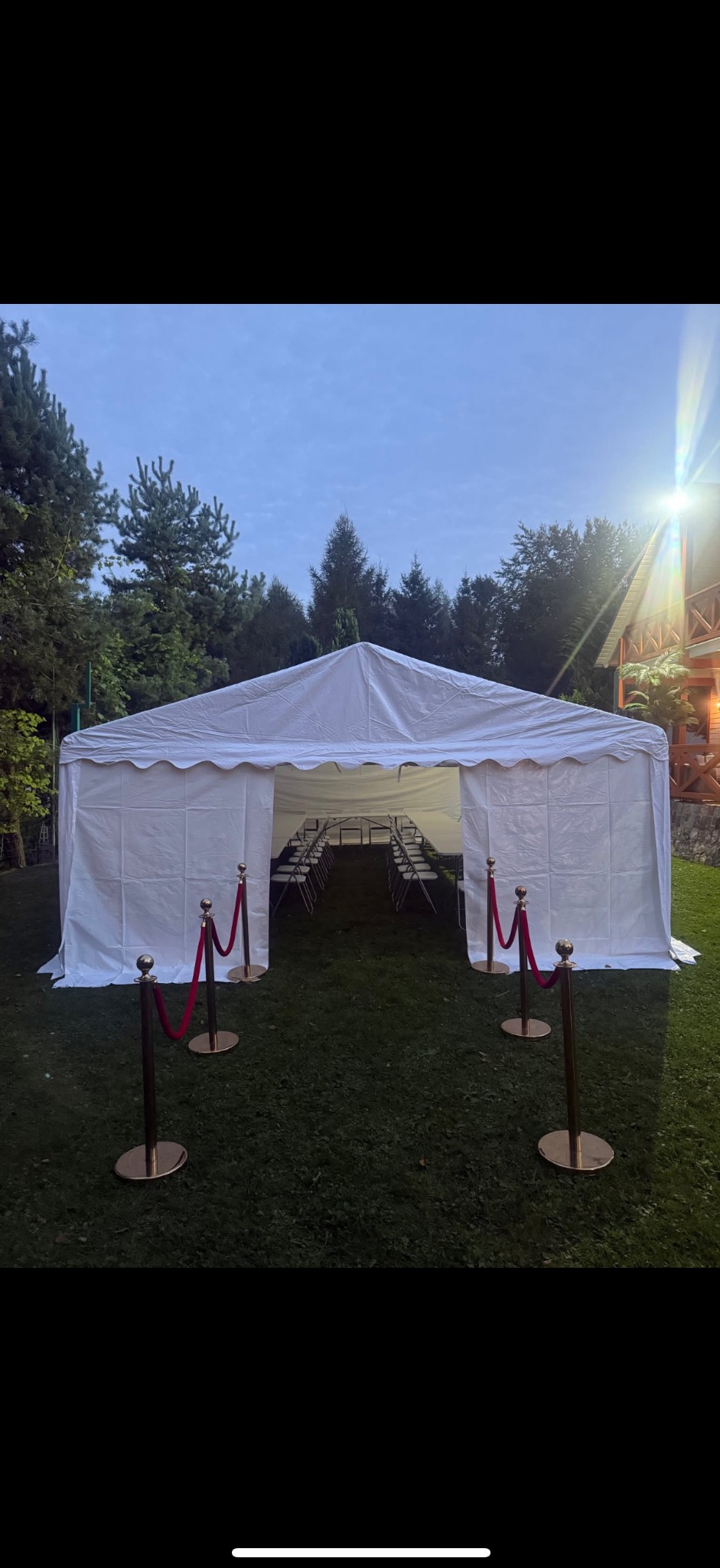Biały namiot eventowy na trawie, z rzędami krzeseł wewnątrz, oddzielony od wejścia złotymi słupkami z czerwonym sznurem. Tło stanowią drzewa i budynek.