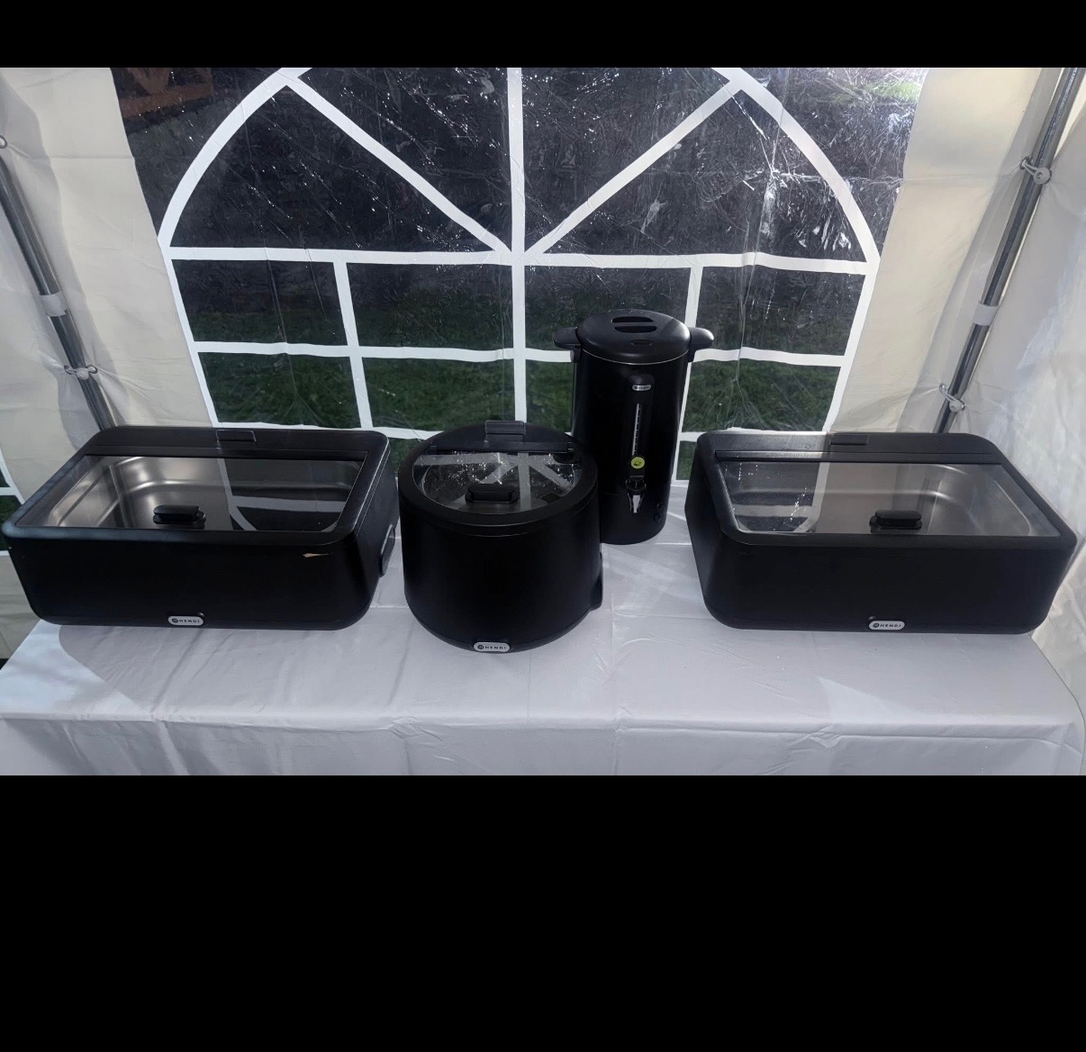 Czarny sprzęt gastronomiczny Hendi: bemar, podgrzewacz do zupy i dozownik napojów na białym obrusie w namiocie. Widok z przodu, oświetlenie naturalne.