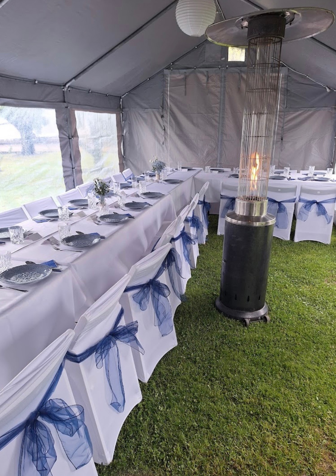 Uroczysty stół pod namiotem, nakryty białą zastawą i dekorowany niebieskimi wstążkami na krzesłach. Widoczny grzejnik gazowy i lampion.