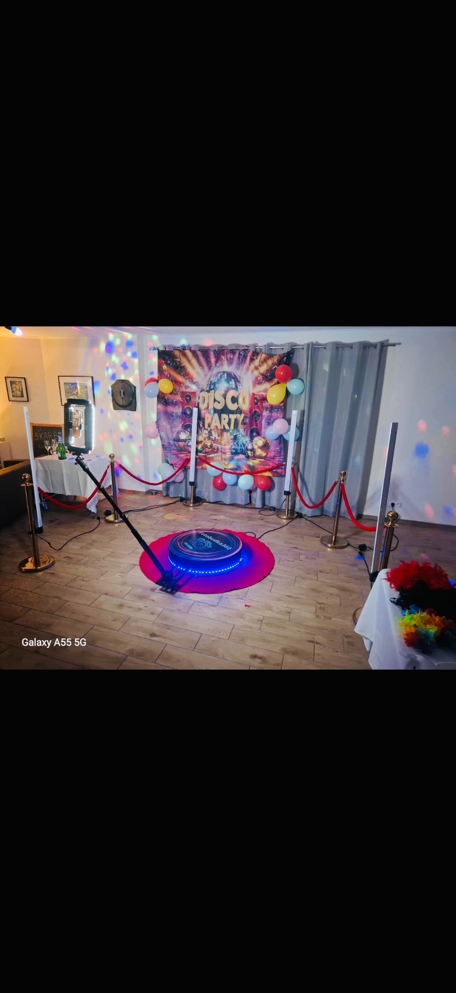 Rozstawiona fotobudka 360 na różowym podeście, otoczona słupkami z czerwonym sznurem. W tle dekoracja 'Disco Party' z balonami i światłami dyskotekowymi.