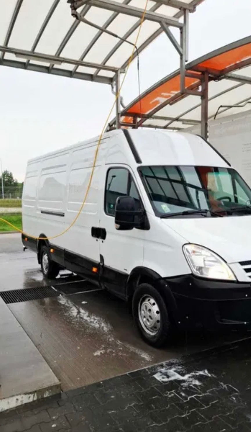 Biały bus w myjni automatycznej, z widoczną lancą do mycia ciśnieniowego i mokrą nawierzchnią. Ujęcie z boku, z lekkiego dołu, pod zadaszeniem myjni.