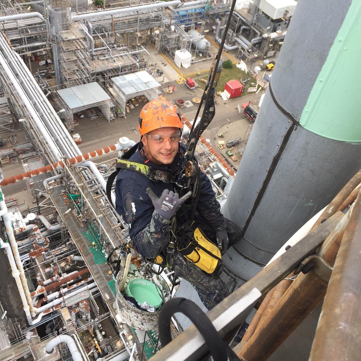 Pracownik w uprzęży, z uśmiechem, na wysokości przemysłowej instalacji. Widok z góry na rozległą infrastrukturę rurociągów i budynków. Kask i okulary ochronne.