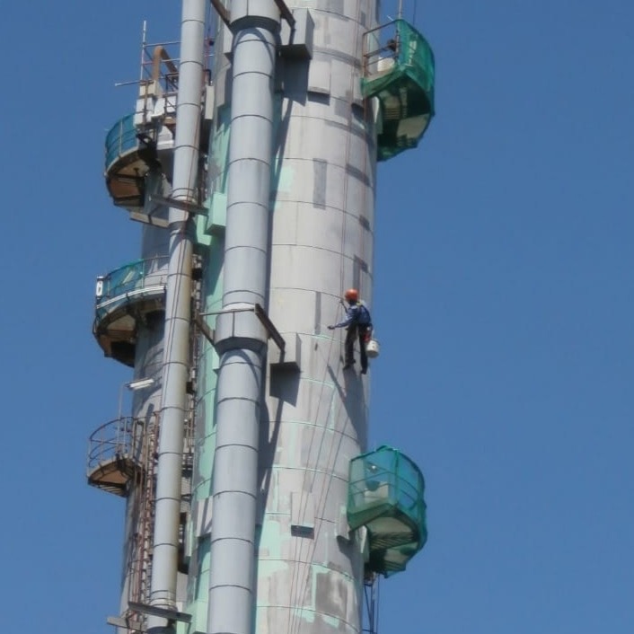 Pracownik w kasku maluje wysoki komin przemysłowy z użyciem technik alpinistycznych. Widoczne rusztowania i instalacje. Błękitne niebo w tle.