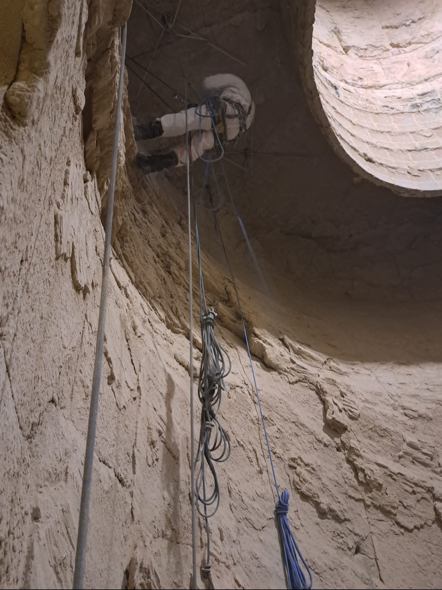 Pracownik w kombinezonie, zabezpieczony linami, wykonuje prace na wysokości wewnątrz studni. Widok z dołu, oświetlenie naturalne.