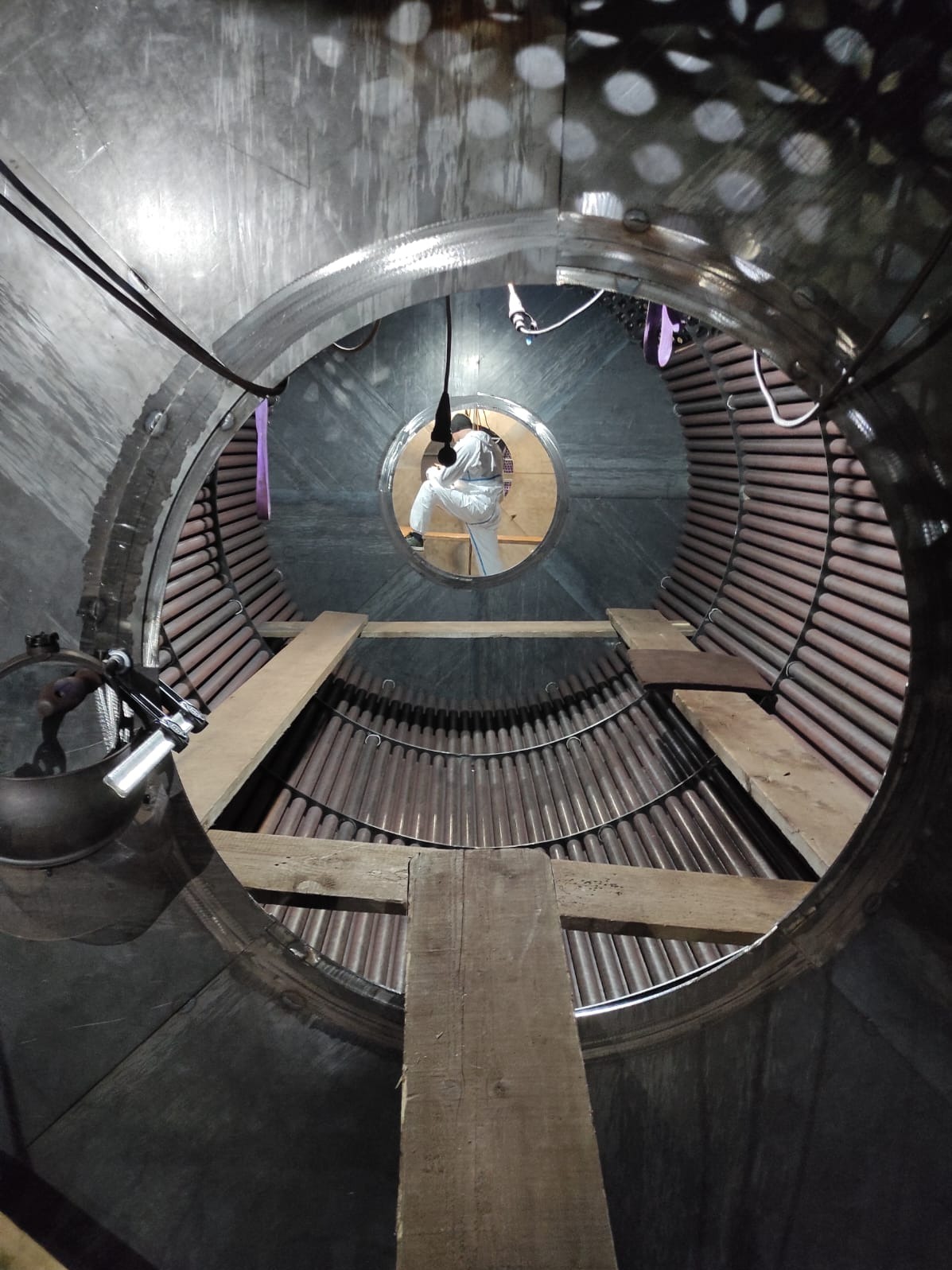 Wnętrze kotła przemysłowego z pracownikiem w kombinezonie ochronnym, widoczne rury i drewniane rusztowania. Nietypowy kadr z perspektywy wnętrza urządzenia.