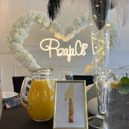 ~ADRIA~ CENTRUM BIZNESOWE JOLANTA ADRIANOWICZ - Dekoracja stołu z numerem '1' w złotej ramce, dzbanki z napojami, kwiatowe serce i pióra. Elegancki wystrój na przyjęcie.