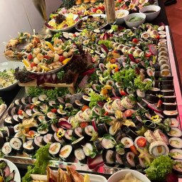 ~ADRIA~ CENTRUM BIZNESOWE JOLANTA ADRIANOWICZ - Bogato zastawiony stół cateringowy z sushi, przekąskami, sałatkami i owocami, udekorowany zielenią i kwiatami, tworzący elegancką kompozycję.