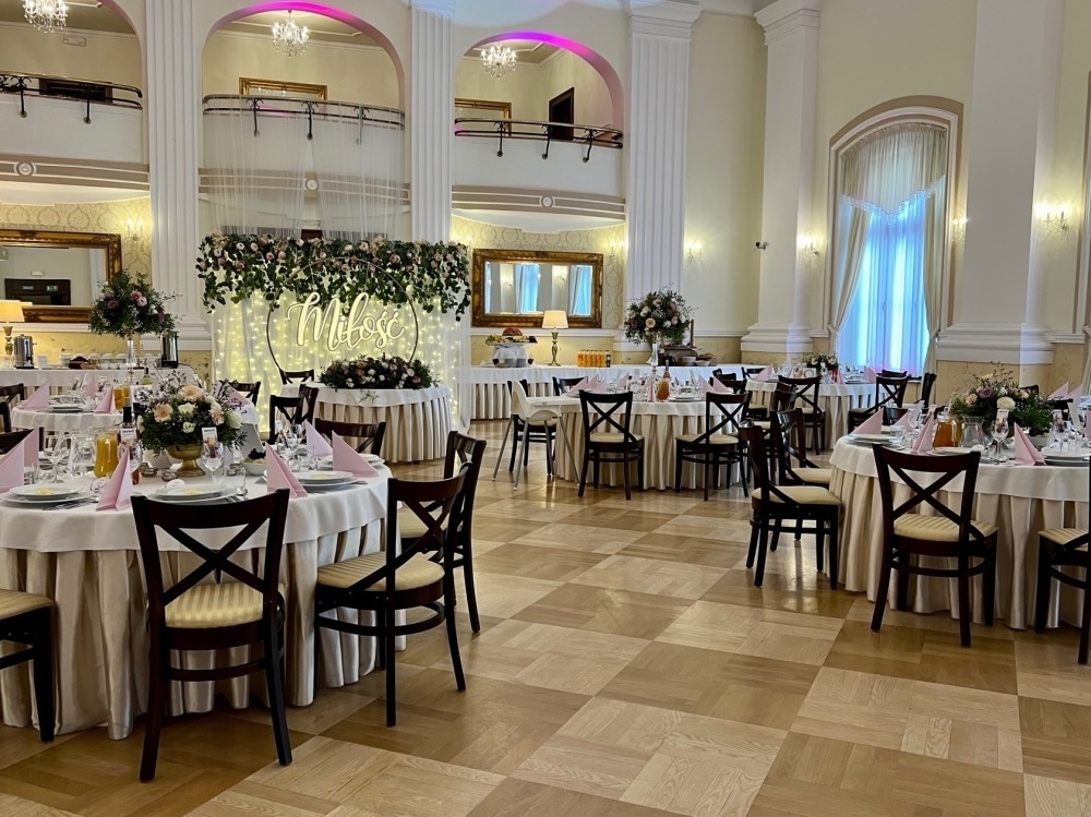 Elegancka sala balowa z okrągłymi stołami, dekoracjami kwiatowymi i napisem 'Miłość' podświetlonym girlandą. Parkiet w szachownicę, kolumny i balkony w tle.
