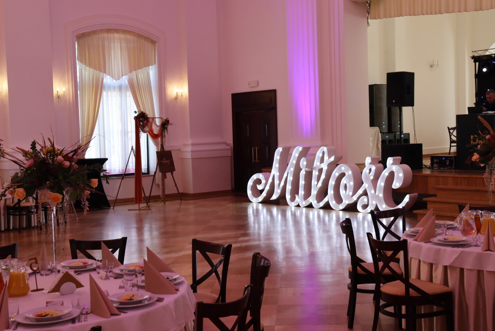 Sala balowa z dekoracją 'Miłość' podświetlaną żarówkami, stoły z nakryciami i kwiatami, scena w tle. Dekoracja sali na wesele lub inną uroczystość.