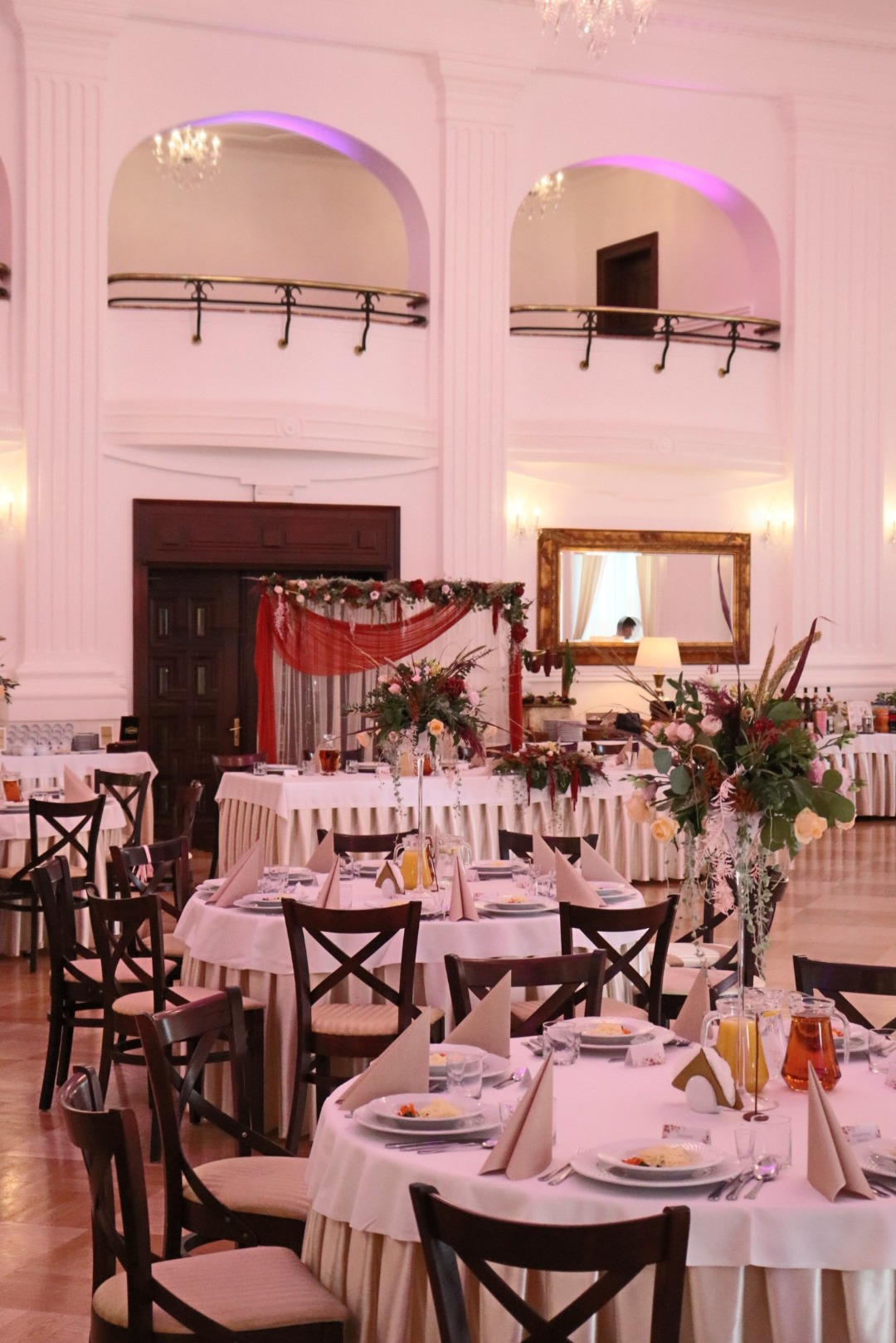 Elegancka sala balowa z okrągłymi stołami, nakryciami i dekoracjami kwiatowymi. W tle kolumny i antresola. Oświetlenie w ciepłych odcieniach.