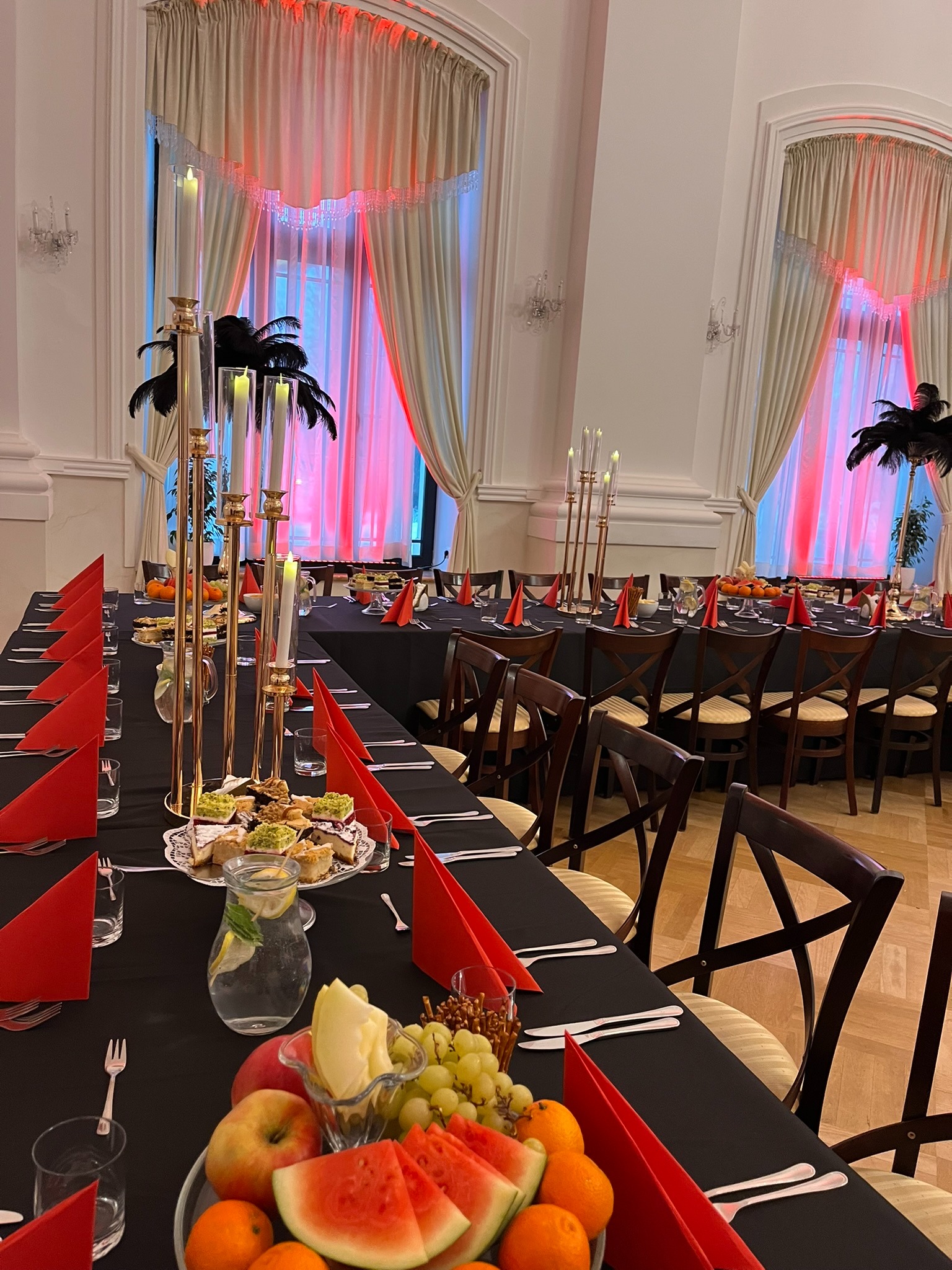 Uroczysta sala balowa z długimi stołami nakrytymi czarnymi obrusami, czerwonymi serwetkami i dekoracjami ze świec i piór. Na stołach owoce i ciasta.