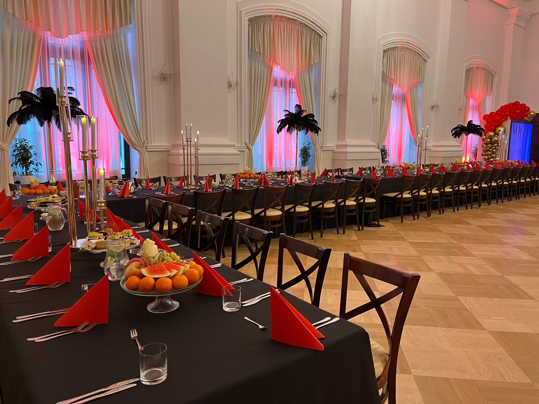 Uroczysta sala balowa z długimi stołami, czarnymi obrusami, czerwonymi serwetkami, owocami i wysokimi świecznikami. Dekoracje z piór i balonów.