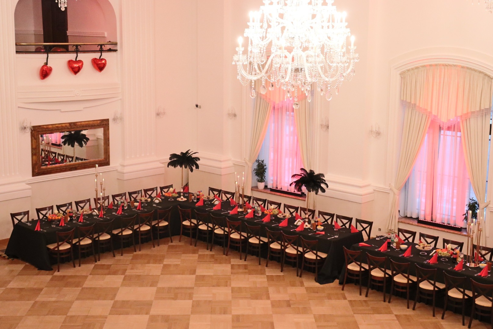 Elegancka sala balowa z długimi stołami nakrytymi czarnymi obrusami, czerwonymi serwetkami i dekoracjami. Kryształowy żyrandol i lustro dodają blasku.