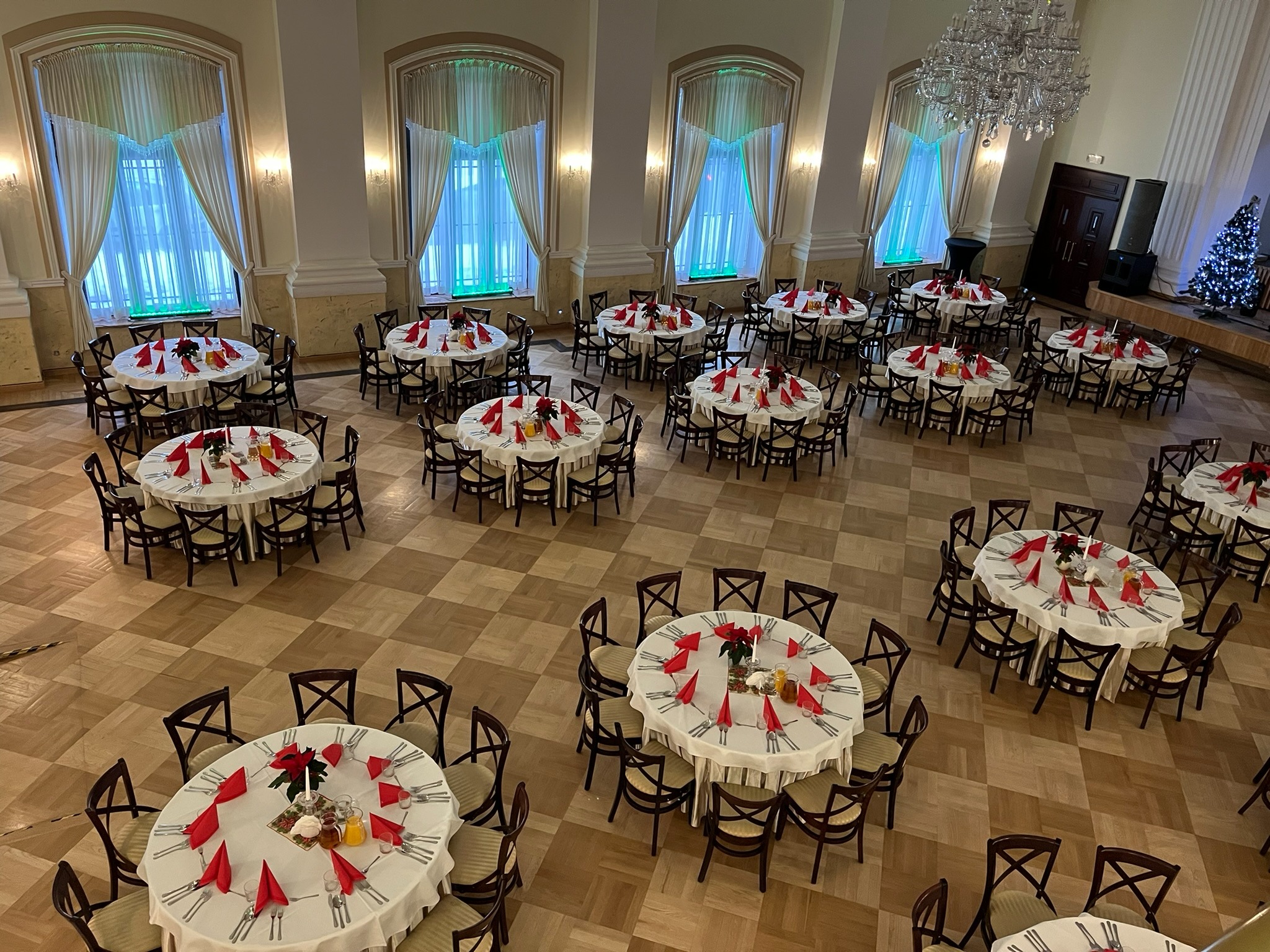 Sala balowa z parkietem, okrągłymi stołami nakrytymi białymi obrusami, czerwonymi serwetkami i kwiatami. W tle okna z białymi zasłonami i choinka.